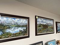 Three framed landscape paintings mounted on wall showcasing mountainous scenes with lakes and rustic cabins.