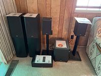 Full speaker setup showing two floorstanding speakers with manuals, two smaller speakers on stands, and a subwoofer with manual and disc on carpet against panel wall.