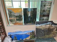 Seven canvas paintings displayed near window and on floor showing mountain, waterfall, lake, forest and urban themes