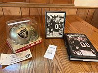 Photo showing silver Raiders mini helmet signed by Jim Otto inside clear plastic case next to framed autographed photo and book