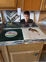 Overview of vinyl record covers including John Lennon, Victory, Christopher Cross, and others arranged on a wooden surface.