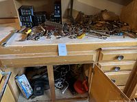 Full view of pile of assorted hand tools and unopened small black boxes on wooden shelf inside cabinet, plus some items stored below.