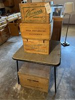 Four wooden crates stacked on a folding card table in a kitchen setting: Vernor's Ginger Ale top crate, High Explosives middle, Nesbitt's Beer lower, and Army Bread crate beneath the table.