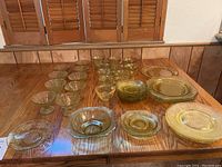 Photo showing multiple yellow Depression glass pieces on a wooden table including plates, bowls, dessert glasses, and small cups.