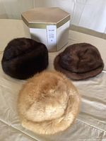 Three mink hats in light, medium, and dark brown colors placed in front of a vintage hexagonal hat box on a beige cloth surface.