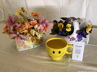 Photo showing two ceramic baskets with artificial floral arrangements and one large yellow smiley cup