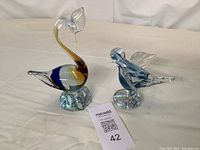 Two glass bird figurines: a larger swan with swirled color glass and a smaller blue and white swirled glass bird, both standing on textured clear glass bases.
