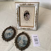 Three ornate picture frames displayed on a white surface: one large rectangular frame with white and gold decoration; two small oval bronze-colored decorative frames with art prints inside.