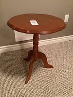 Front view of a medium brown maple piecrust occasional table with scalloped edge top and turned pedestal base ending in three curved legs. Table stands on carpet against wall.