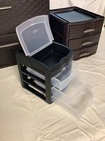 View of the three plastic storage drawer boxes including the black unit with clear drawers and top lid open, plus two dark brown or black units with three drawers and silver handles