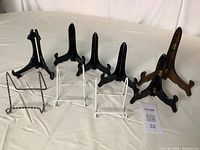 Seven assorted plate stands displayed on a white surface including black plastic, white metal, wood, and metal wire stands.