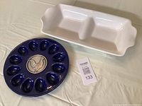 Blue ceramic deviled egg tray with twelve egg-shaped sections and central chicken design alongside a white divided serving tray with two compartments and decorative handles.