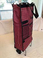 Collapsible shopping bag standing upright showing side view with pinstriped red fabric, wheels, and metal legs.