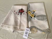 Two folded table toppers on a white surface: one sheer round topper with embroidered red roses folded on left, one square topper with yellow embroidered flowers and cut-outs folded on right.