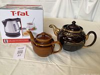 T-fal electric kettle in its box alongside two vintage teapots, black and gold teapot and smaller brown teapot