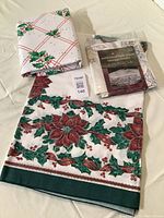 Top view of the three folded table linens including floral poinsettia cotton tablecloth, plastic flannel-backed tablecloth with holly design, and packaged embroidered table topper.