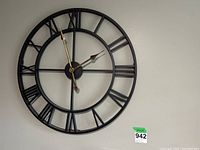 Front view of black metal wall clock with Roman numerals and gold-tone hands on white wall, measuring 24x24 inches.