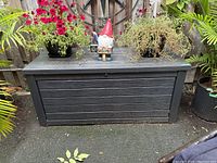 Black resin deck box outside with plants and a garden gnome on top, showcasing overall size and condition.