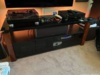 Front view of media stand with items on top including turntable, remote controls, and electronic devices. Stand has four glass front doors and wood veneer sides.