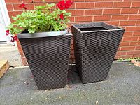 Two outdoor dark brown faux basket weave resin planters side by side; one contains a red geranium plant with green leaves, the other is empty