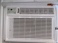 Front view of white portable window air conditioner installed in window frame showing vent grills and control panel with buttons and lights.