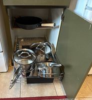 Photo of four stainless steel Ingenia pots with lids and one coated metal wok with wood handle stored inside an open kitchen cabinet.
