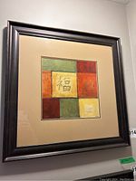 Framed abstract print featuring a square composition of colorful blocks and central Asian character hung on a white wall.