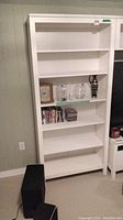 Full view of solid white painted wood shelf unit with 6 levels, some items on middle shelves for scale, plain wall background