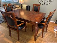 Front and side angle view of wood dining table with four matching chairs around it. Table top shows wood veneer inlay pattern. Chairs have black cushioned oval backs and seats.