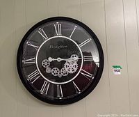 Front view of Westzytturm round black wall clock with white Roman numerals and visible moving silver gears at center.