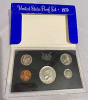 1970 US Proof Coin Set housed in original blue-white cardboard sleeve. View of set inside clear plastic case with black backing showing five proof coins.