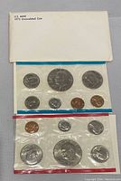 Two complete 1973 US Mint uncirculated coin sets in original cellophane packaging. One set with blue trim, one with red trim. Each set contains 5 coins including penny, nickel, dime, quarter, and half dollar.