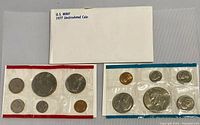 Photo showing two sealed US Mint coin sets from 1977 with one in red (Denver) and one in blue (Philadelphia) cellophane packaging, plus the original envelope.