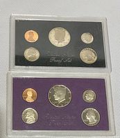 Both 1983 and 1987 US proof coin sets displayed together, showing front of sealed plastic cases and coins inside.