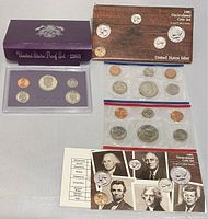 Photo showing both coin sets with their original packaging and accompanying documentation, including boxes for the proof set and uncirculated mint set.