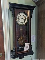 Full view of antique wooden regulator wall clock mounted on wall, showing wooden case, glass front, pendulum, and dial with Roman numerals.
