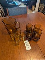 Overview of the lot showing amber glass pitcher, 4 diamond pattern glasses, and 4 heavy brutalist amber goblets on wooden table