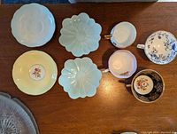 Top-down photo showing all 4 orphan tea cups and 4 saucers placed on a wooden surface, showcasing various colors and patterns including light blue flower-shaped saucers, yellow saucer with floral center, and different floral cups.