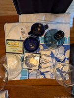 Overview photo showing blue floral tablecloth, blue ceramic sugar and creamer set, clear blue glass decanter, four white pig napkin rings, coasters, vases, corn holders in packaging.