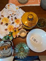 Overall view showing white tablecloth with floral pattern, white bowl, mustard yellow ceramic coffee jar with lid, Mikasa ceramic creamer, deer wood salt and pepper shakers, duck wooden napkin rings, and a green lace glass bowl with scalloped edge on wooden surface.