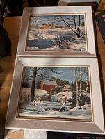 Pair of framed vintage paint by numbers winter landscape paintings laid on a table, showing snow, trees, barns, and fences.