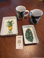 Four mugs and two rectangular serving dishes on wooden table, mugs and dishes with botanical designs.