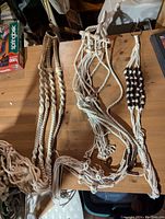 Three macrame plant holders laid on a wooden surface, two long beige rope holders with wood beads and one shorter white string holder with brown wood beads.