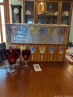 Two boxed sets of Vintage VMC Reims French Harlequin crystal wine glasses in multiple colors, plus four ruby red Cristal D'Arques Durand wine glasses displayed on wooden table matching the title and description.