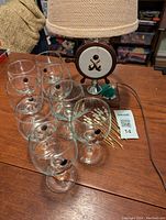 Eight clear glass wine glasses with bowling ball and pin design, arranged on a wooden surface with the lamp and picks present in background.