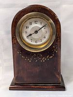 Front view of the antique wooden tombstone style alarm clock with decorated lower half of the face surround showing clock hands and numerals.