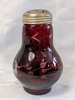 Front view of cranberry glass sugar shaker showing carved floral and leaf motifs and metal shaker lid.