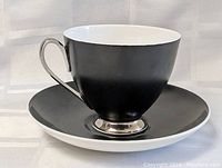 Close-up of one cup and saucer set showing black exterior cup, white interior, silver metal handle and base, and black saucer.