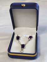 Necklace displayed inside blue and gold jewelry box, showing three square amethyst stones and clover charm.