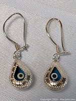 Pair of silver dangle earrings shaped as teardrops with blue and white enamel evil eye motifs.
